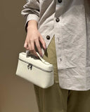 White vegan leather crossbody handbag with silver zipper and logo emblem held by person in beige shirt and olive pants