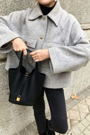 Woman holding a black vegan leather bucket bag with gold lock detail, wearing a gray jacket and black pants outdoors