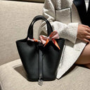 Black vegan leather bucket bag with gold lock and decorative scarf on couch with person in white coat