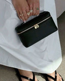 Black vegan leather crossbody handbag with silver logo and zip detail held by a woman in white dress