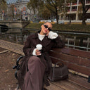 woman sitting on bench holding coffee cup with black Aveline handbag beside her outdoors