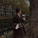 Woman wearing a dark trench coat holding a black Aveline handbag and a coffee cup by a riverside in autumn