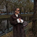 woman wearing dark brown suede trench coat carrying black structured Aveline handbag with adjustable strap near canal in urban autumn setting