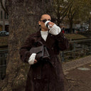 Woman wearing a brown suede trench coat holding a black Aveline handbag and drinking coffee outdoors