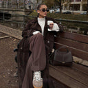 Elegant ebony Aveline handbag in vegan leather with adjustable strap, placed on bench next to woman in suede trench coat.