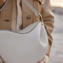 Toscana minimal curved white leather cross body bag with gold hardware and adjustable strap worn on tan coat