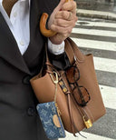 Brown premium vegan leather Piana bucket bag with golden hardware and signature lock, styled with a black blazer and white shirt on city street background