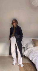 woman wearing Toscana cross body bag in black coat and white pants standing in bedroom interior